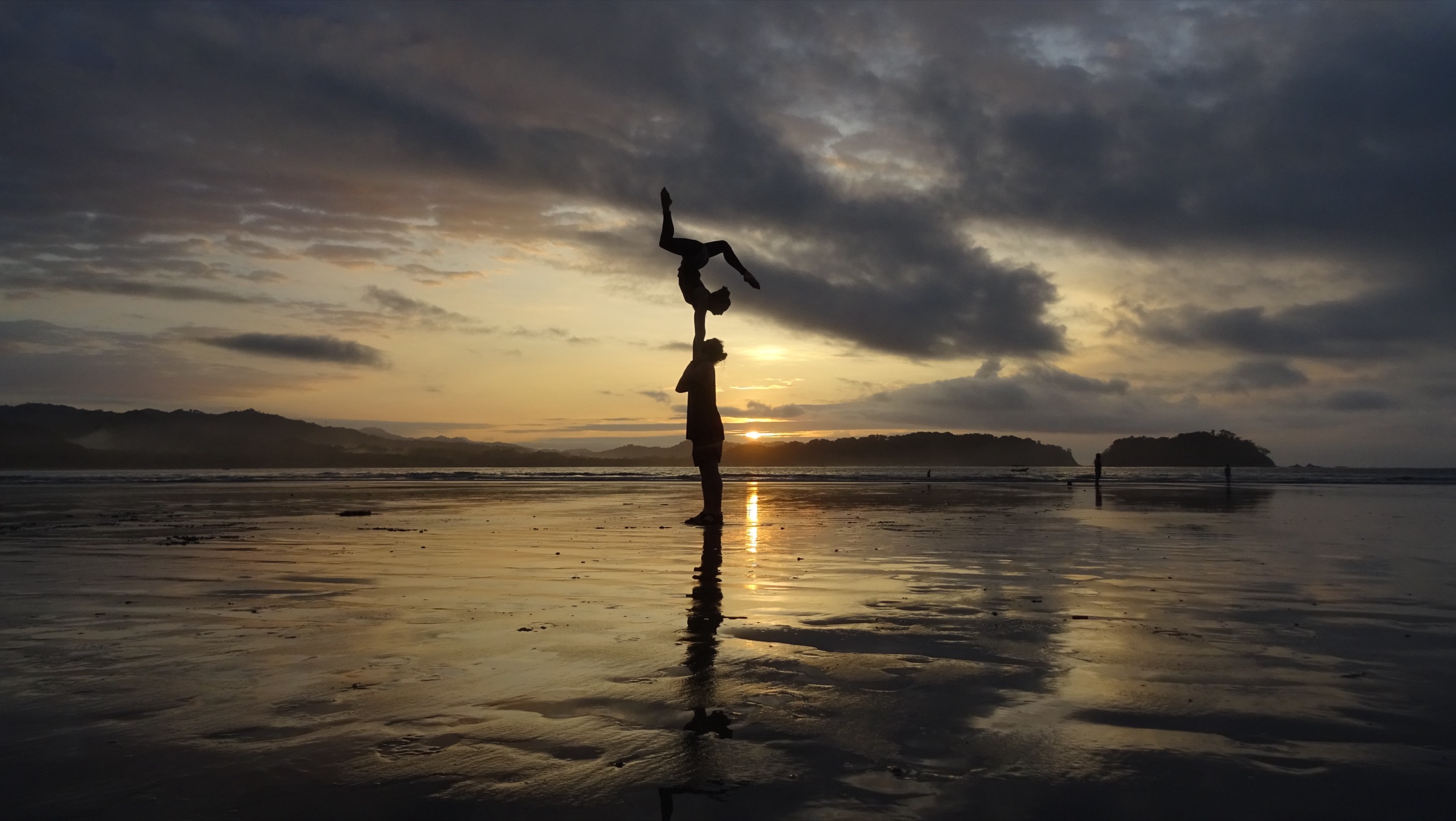Silhouette of Cedric holding a partner overhead on one arm on a mirror-wet beach at sunset