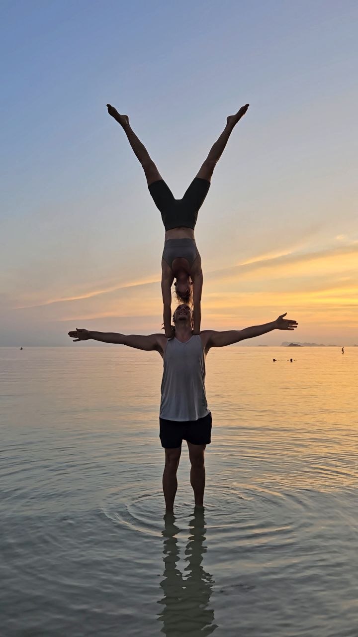 Cedric standing in the sea at sunset, balancing a partner in a handstand on his shoulders